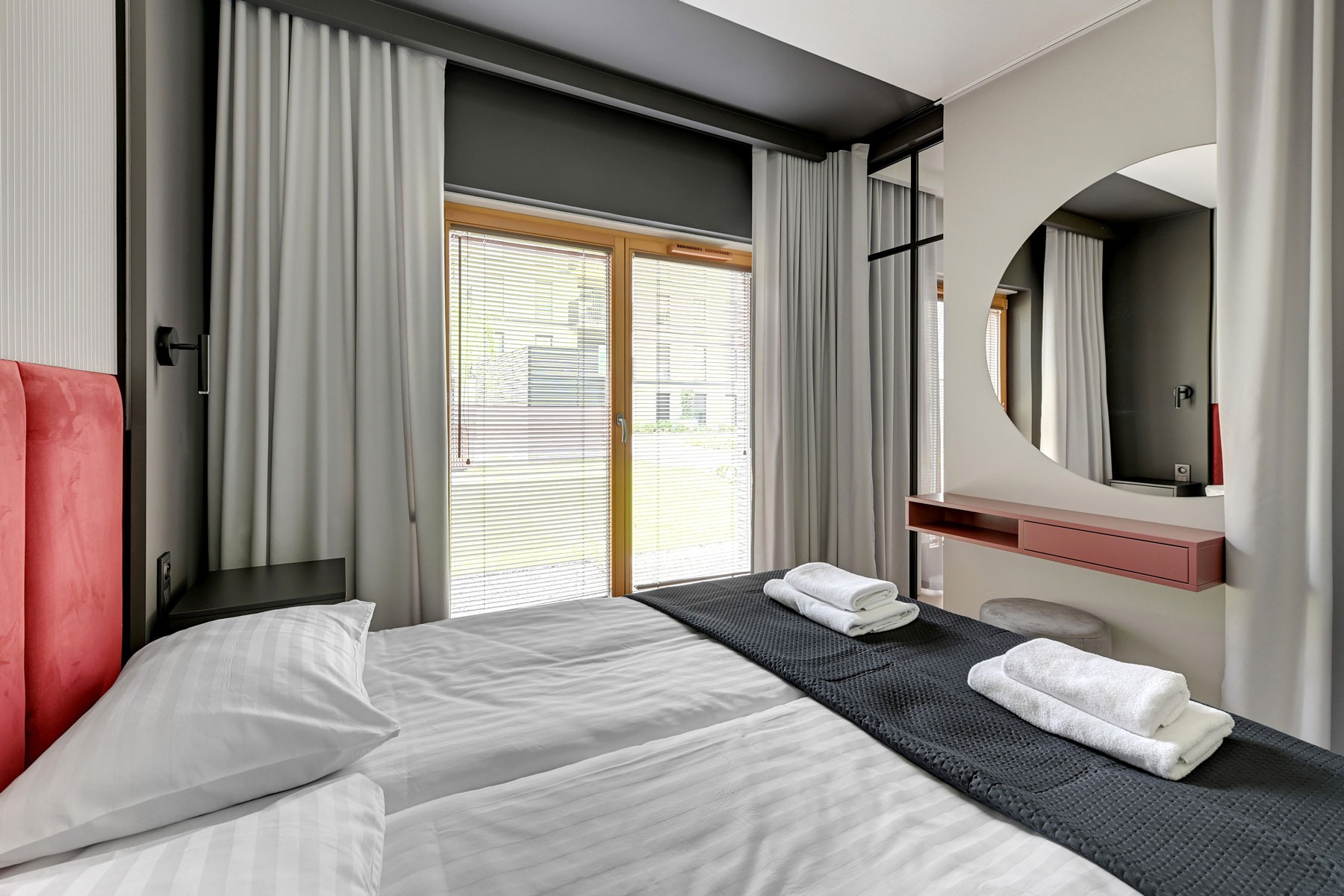 Nowoczesna sypialnia z łóżkiem dwuosobowym, pościel w paski, czerwone wezgłowie, okrągłe lustro nad toaletką z szufladą, szare zasłony i rolety w oknie.