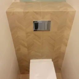 Podtynkowa toaleta z chromowanym przyciskiem spłukującym, obudowana płytkami imitującymi drewno ułożonymi we wz&oacute;r jodełki, widoczne niedokończone wykończenie ściany nad obudową.