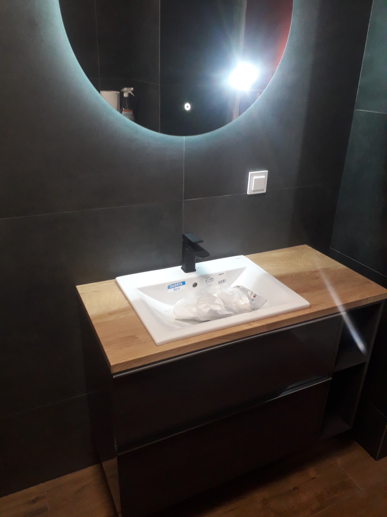 Nowoczesna łazienka z czarnymi płytkami, umywalką na szafce z drewnianym blatem i okrągłym lustrem z podświetleniem LED, widoczny fragment podłogi z paneli.