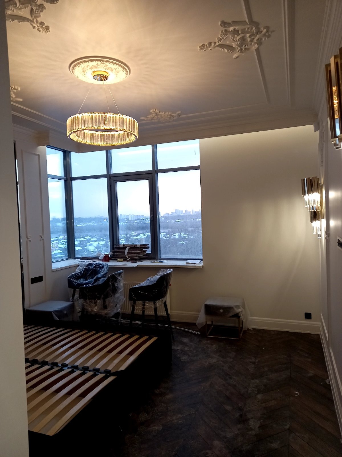 Pokój w trakcie wykańczania: parkiet w jodełkę, niezabezpieczone meble, okno z widokiem na miasto, ozdobny sufit z żyrandolem i kinkietami.