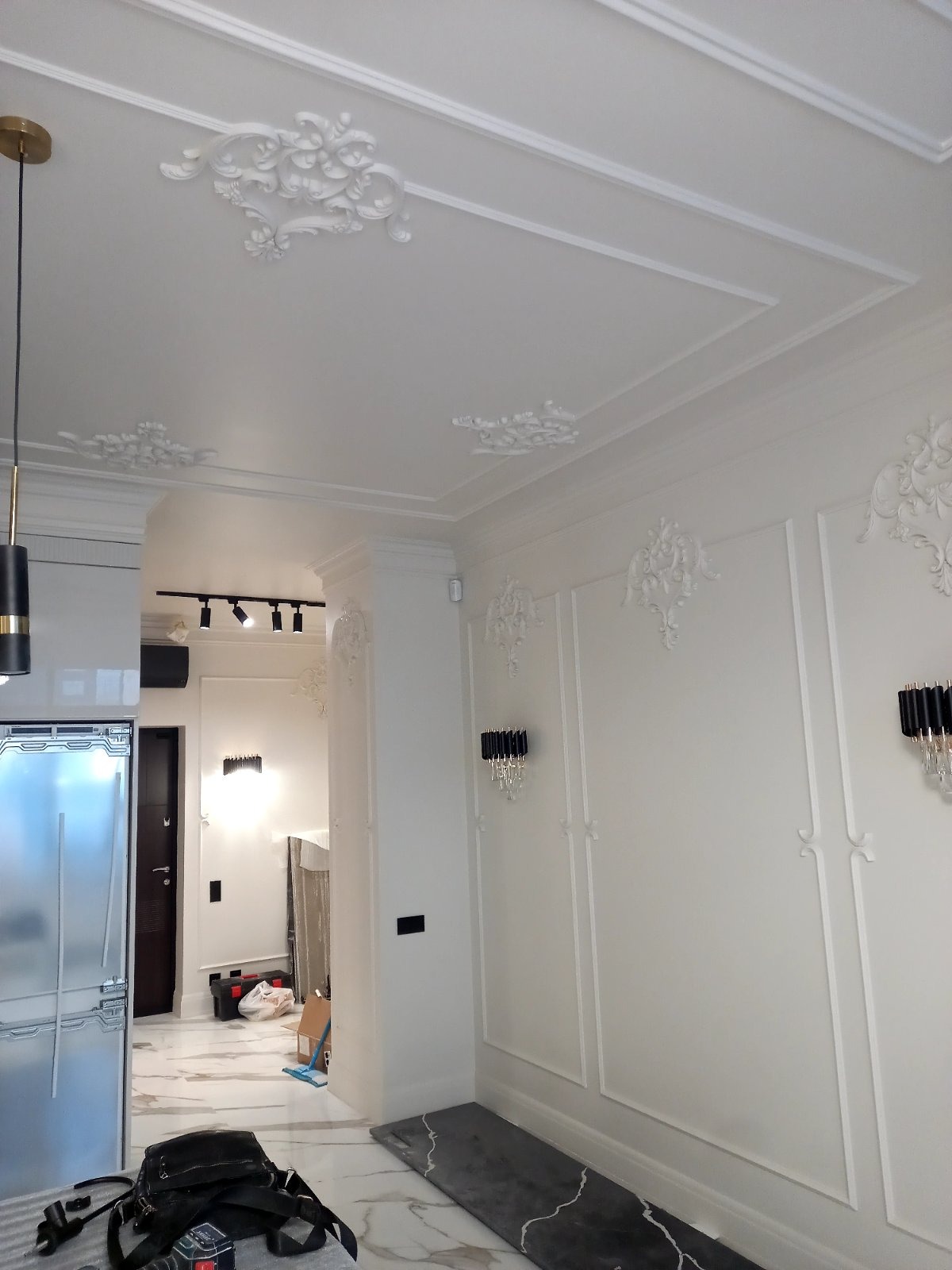 Eleganckie wykończenie wnętrza: białe ściany z dekoracyjnymi listwami i rozetami, marmurowa podłoga, nowoczesne oświetlenie, w tle widoczny fragment kuchni i przedpokoju.