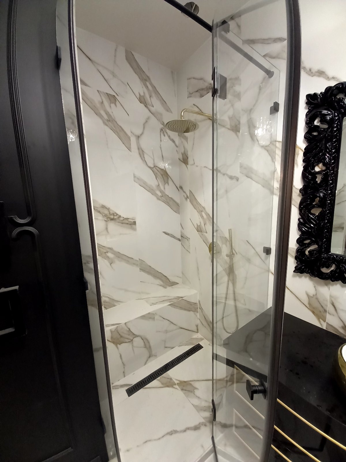 Nowoczesna kabina prysznicowa z marmurowymi płytkami, złotą deszczownicą i czarnym odpływem liniowym, uzupełniona o czarne elementy armatury i ozdobne lustro w ramie.