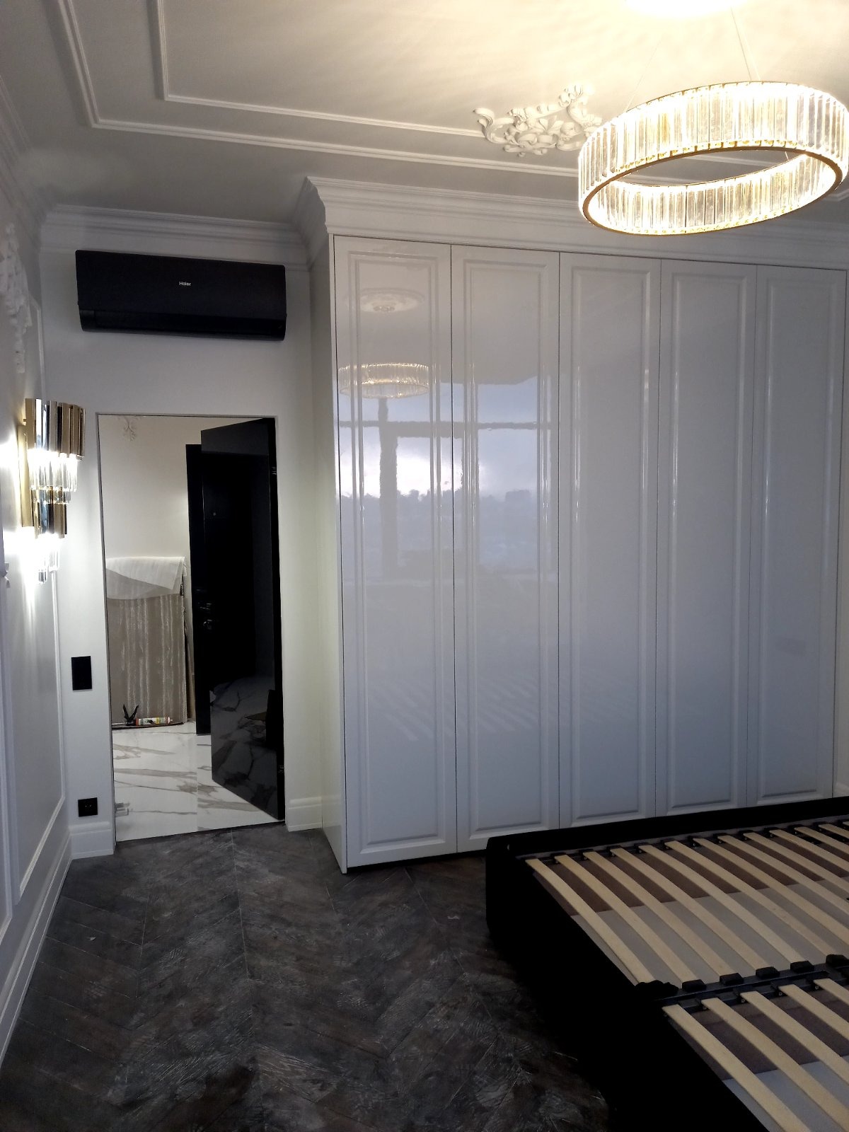 Nowoczesna sypialnia z lśniącą białą szafą w zabudowie, ozdobnym oświetleniem sufitowym i otwartym przejściem do łazienki z marmurową posadzką.