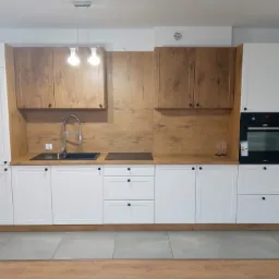 Nowoczesna kuchnia z białymi szafkami dolnymi i drewnopodobnymi szafkami górnymi, czarny zlew i płyta grzewcza, oświetlenie sufitowe z trzema żarówkami.