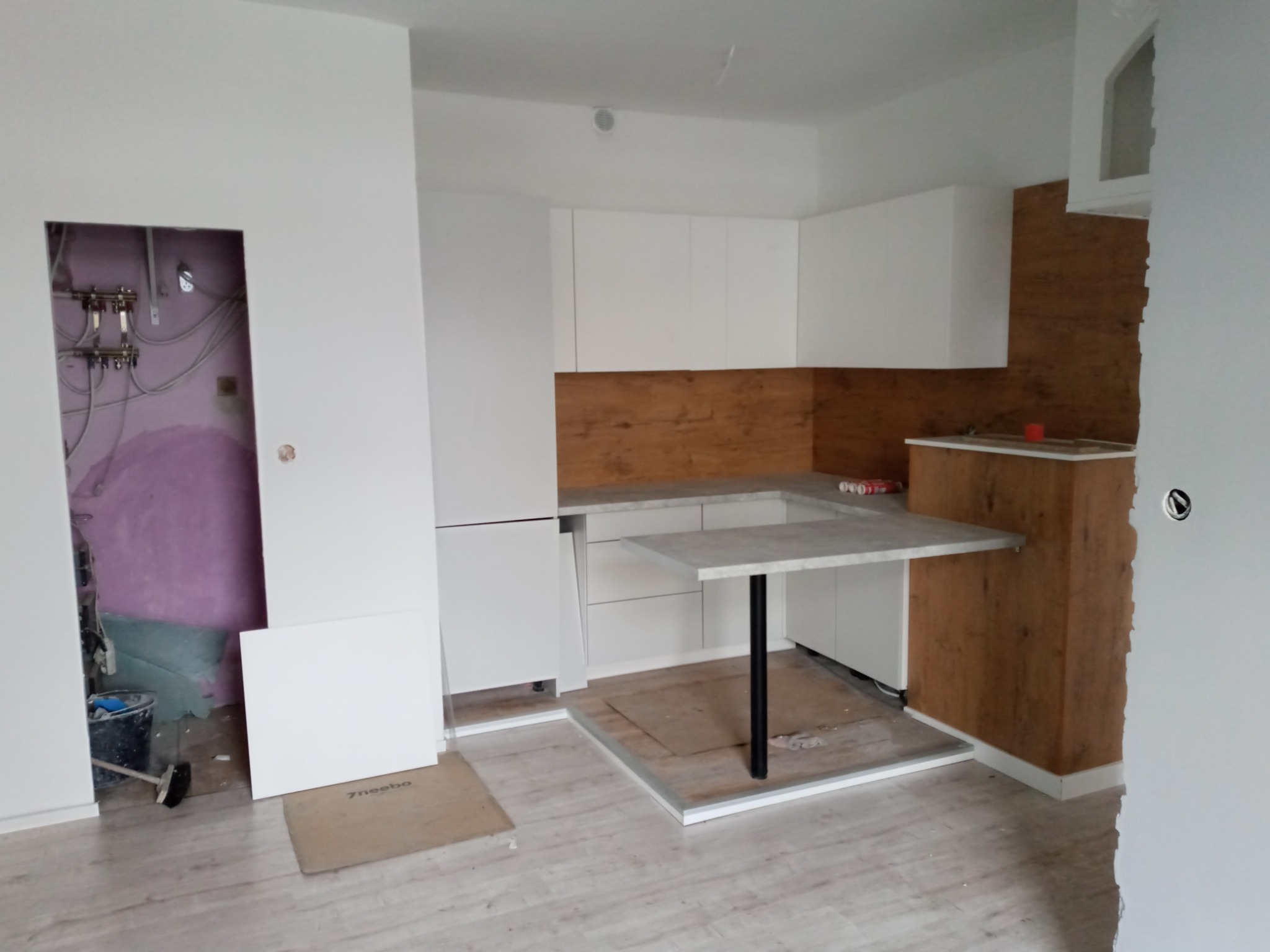 Wnętrze w trakcie montażu: białe szafki kuchenne z drewnopodobnym panelem, wysuwany blat, widoczne niedokończone wykończenia ścian i podłogi, instalacja hydrauliczna w otwartym przejściu.