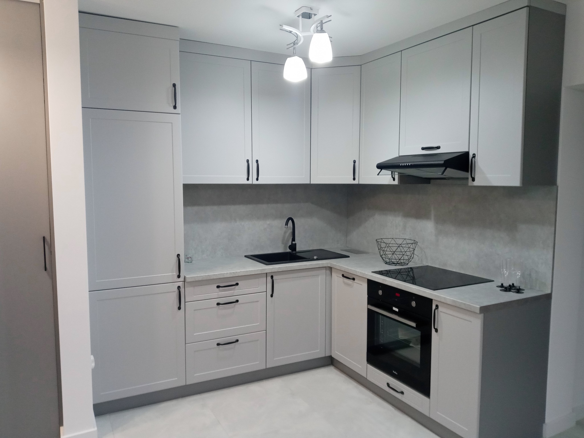 Jasnoszara, narożna zabudowa kuchenna z czarnymi akcentami: bateria, zlew, okap i płyta indukcyjna, oświetlona nowoczesną lampą sufitową.