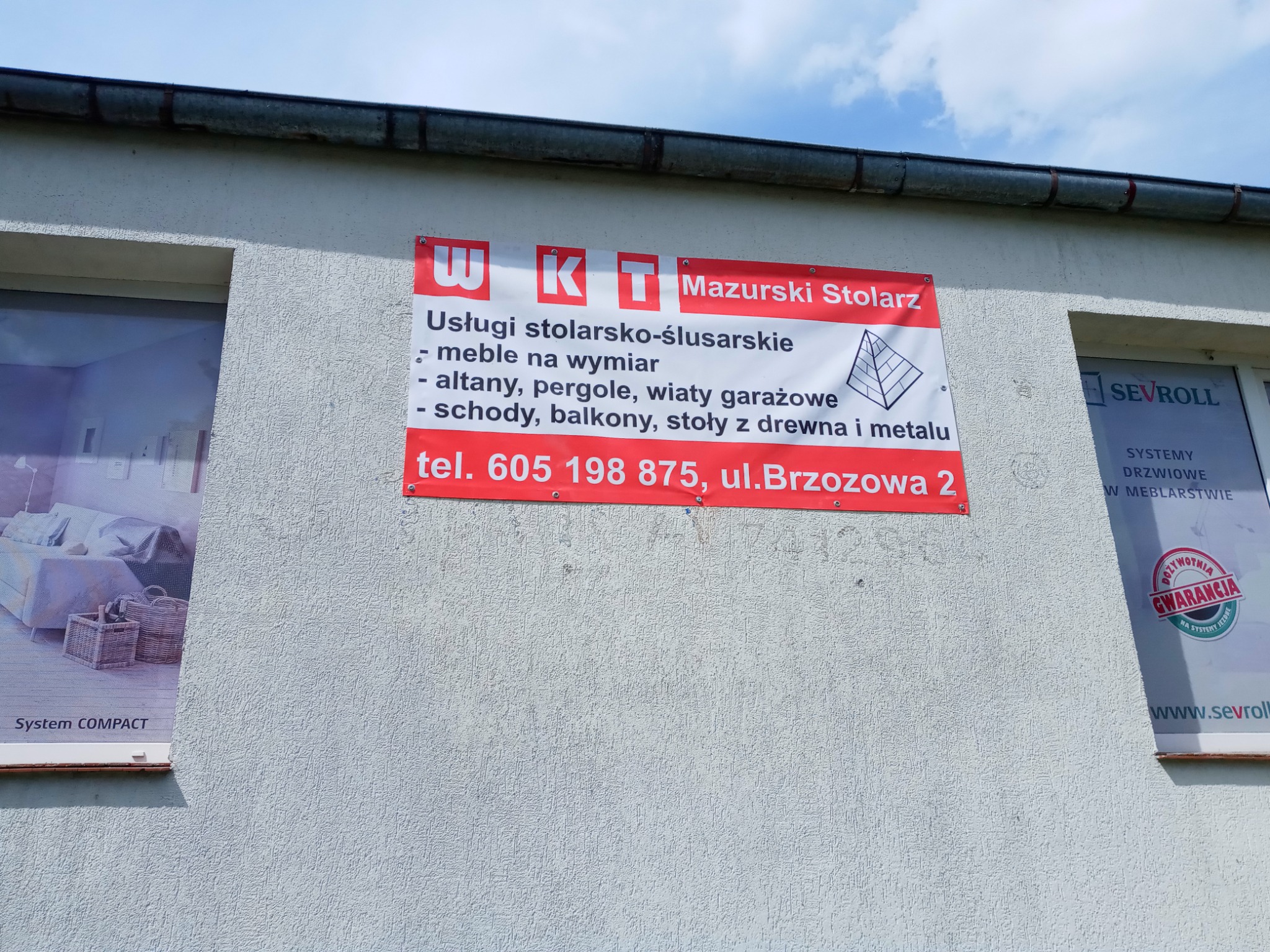 Tablica reklamowa firmy stolarskiej z Mrągowa, oferującej meble na wymiar, altany, pergole i inne konstrukcje z drewna i metalu, umieszczona na ścianie budynku.