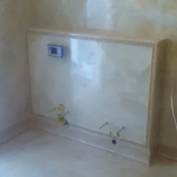 Obudowa stelaża podtynkowego WC z kremowych płytek ceramicznych z dekoracyjnym obramowaniem, widoczne przyłącza wodne i kanalizacyjne, na podłodze płytki w odcieniach beżu.