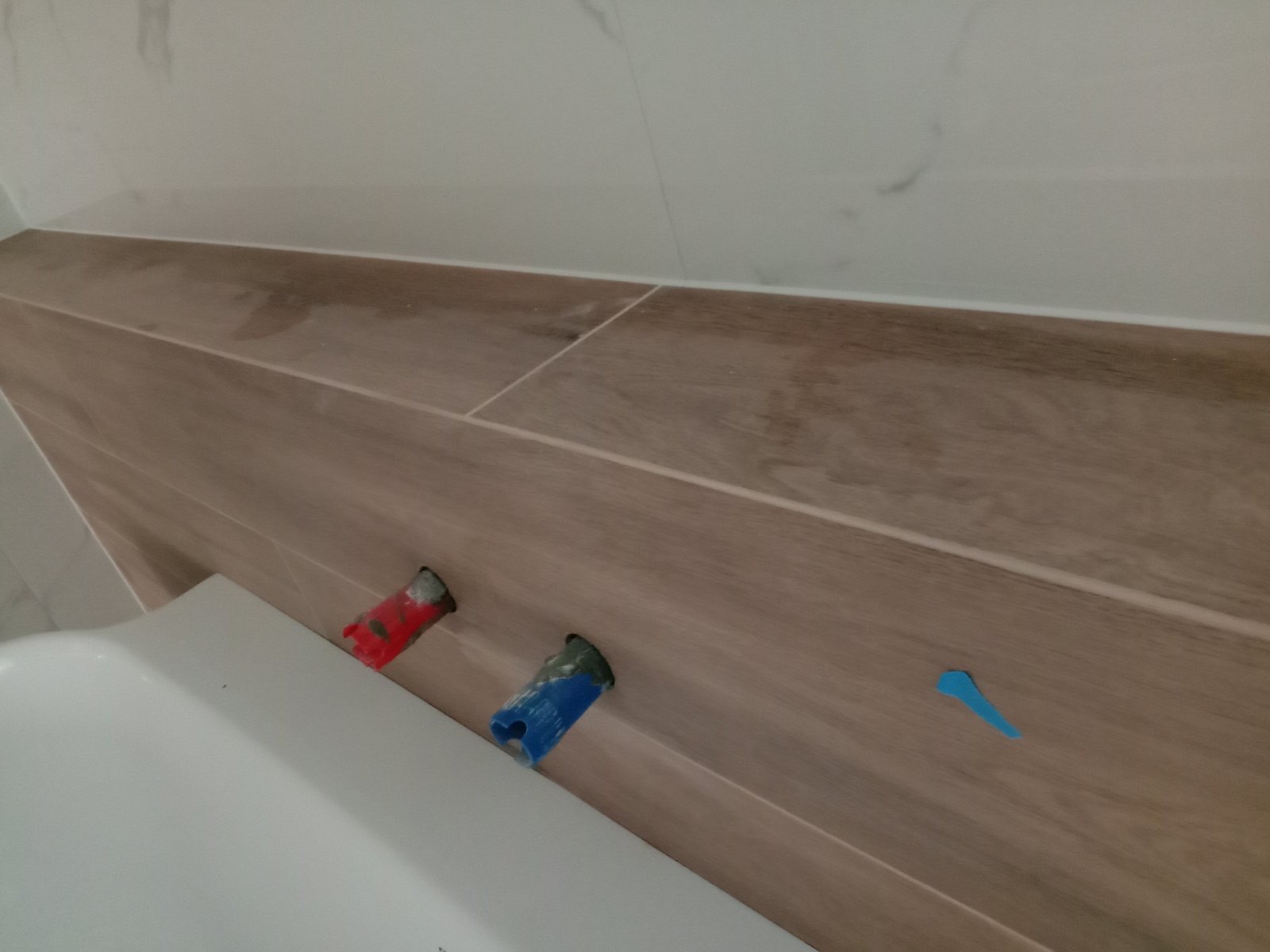 Fragment łazienki po ułożeniu płytek imitujących drewno, widoczne wystające rury z zaślepkami w kolorze czerwonym i niebieskim oraz fragment wanny.