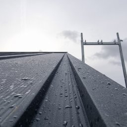 Dom Drzewa - Metalowe pokrycie dachowe w kroplach deszczu, sfotografowane pod kątem, z widocznym fragmentem rusztowania i pochmurnym niebem w tle.