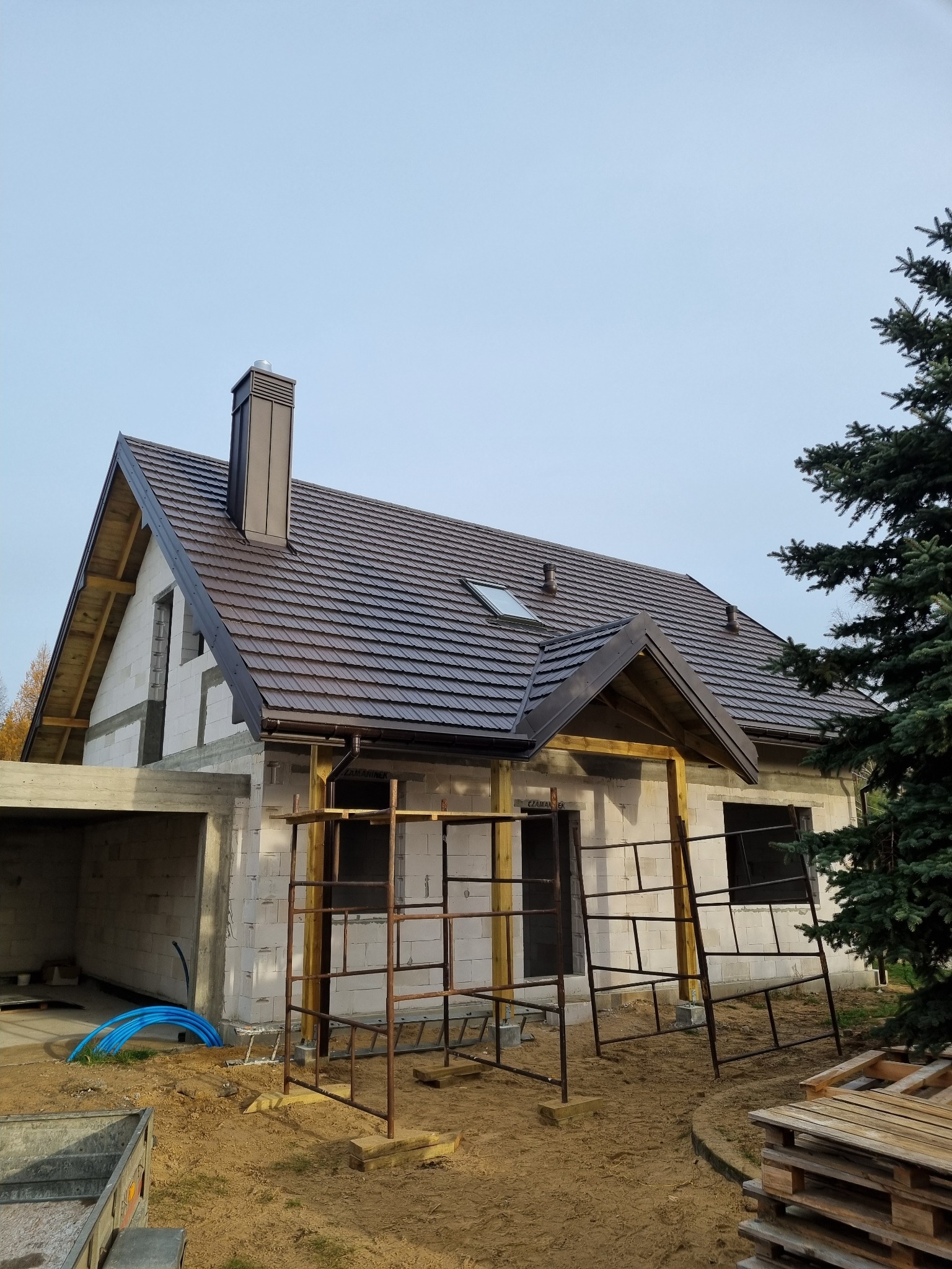 Dom jednorodzinny w trakcie budowy z brązowym dachem z dachówki ceramicznej, widoczne rusztowania i nieocieplone ściany z bloczków.