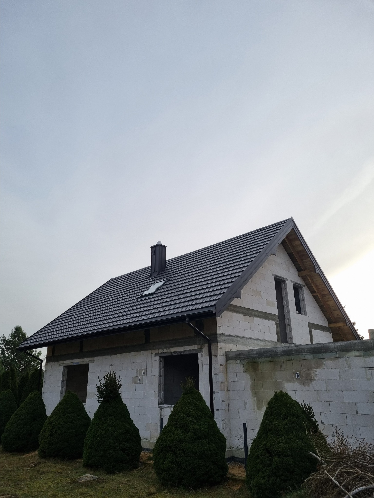 Dom w trakcie budowy z nowym, ciemnoszarym pokryciem dachowym, widoczny komin i okno dachowe, ściany z białego bloczka, ozdobne krzewy iglaste przed budynkiem.
