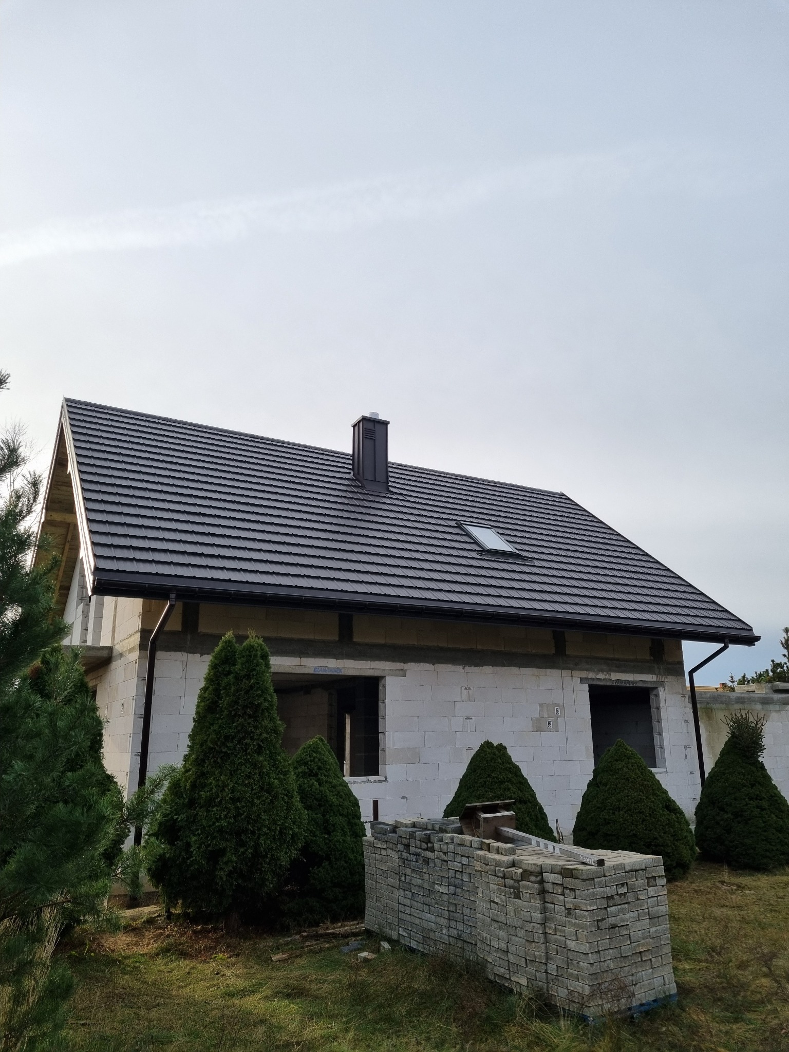 Dom jednorodzinny w Płocku z nowym, ciemnoszarym pokryciem dachowym z blachodachówki, widoczny komin i okno dachowe, budynek w stanie surowym zamkniętym z oknami bez szyb, na pierwszym planie...