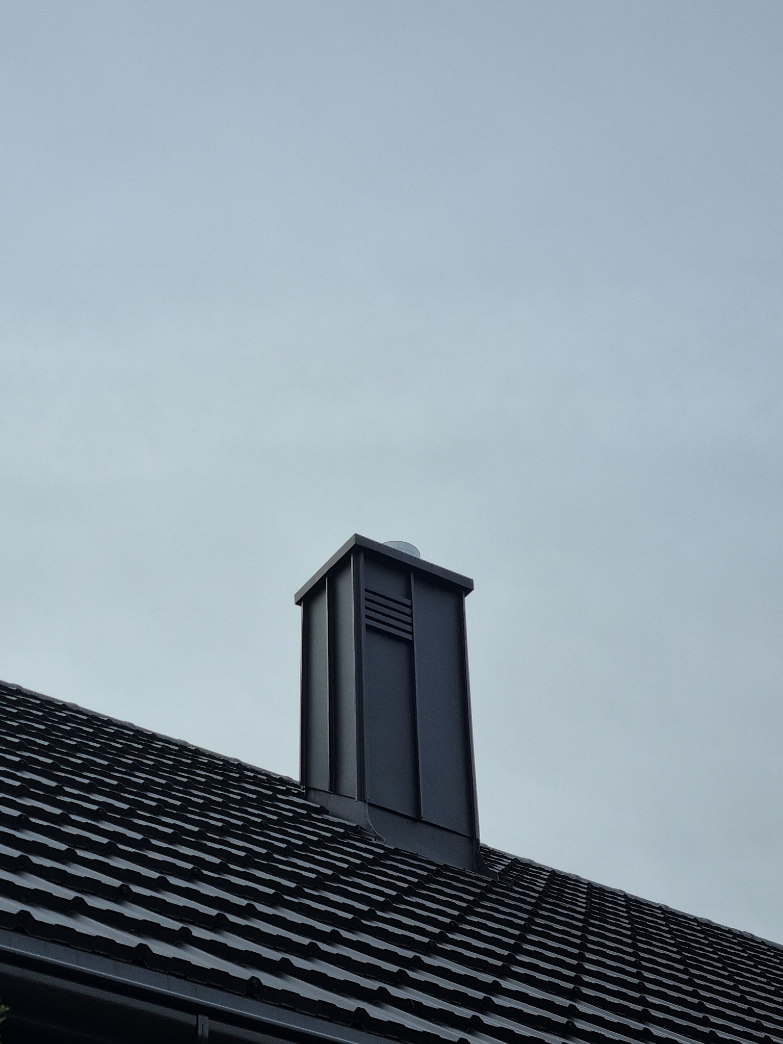 Nowoczesny komin wentylacyjny w kolorze grafitowym, wystający z ciemnego dachu pokrytego dachówką ceramiczną na tle pochmurnego nieba.