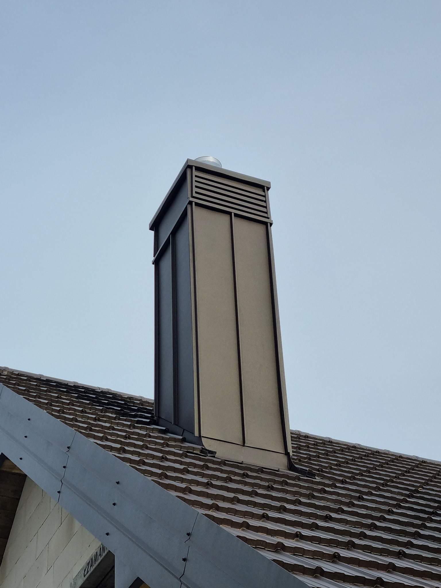 Nowy, prostokątny komin z blachy w kolorze szaro-brązowym, wystający z dachu pokrytego brązową dachówką ceramiczną, widziany z dołu na tle jasnego nieba.