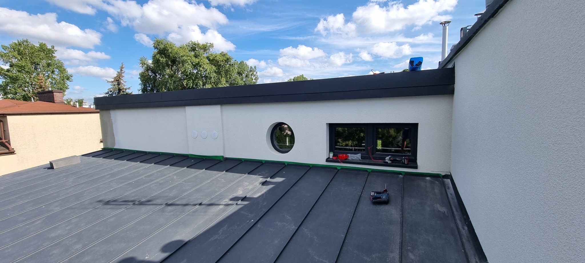 Nowoczesny dach z blachy na rąbek stojący w kolorze grafitowym, z oknem i okrągłym bulajem w białej elewacji, narzędzia na dachu, widok na błękitne niebo z chmurami.
