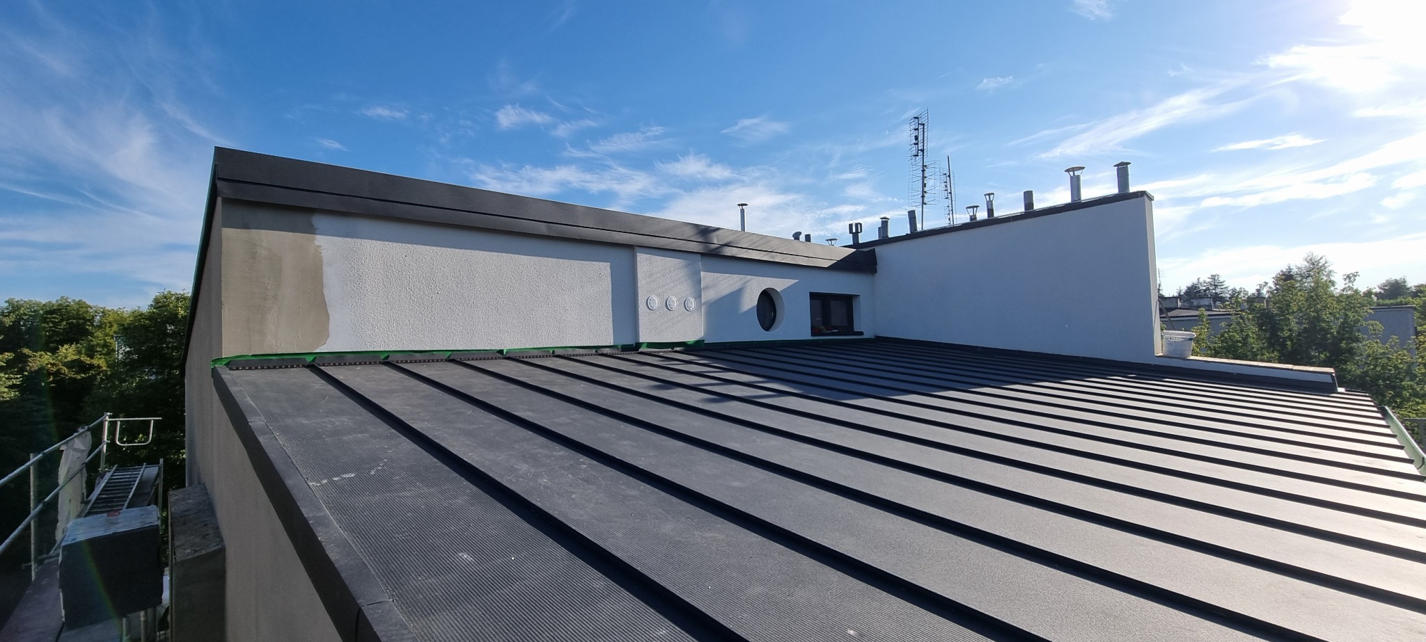 Nowoczesny dach płaski z czarnej blachy na budynku z białą elewacją, widoczne kominy, antena i okrągłe okno.