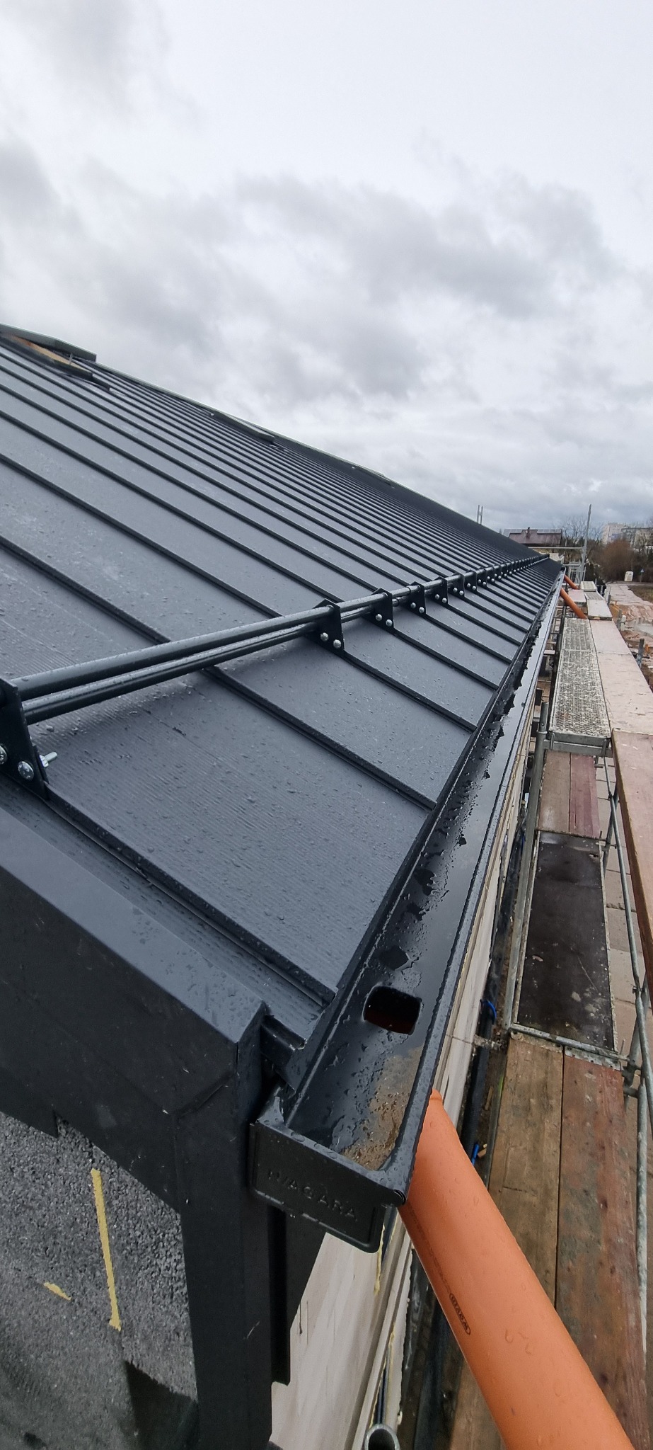 Nowoczesny, ciemnoszary dach z blachy na rąbek stojący, widziany z perspektywy rusztowania, z zamontowanym systemem orynnowania z rurą spustową w kolorze pomarańczowym i uchwytami przeciwśniegowymi...