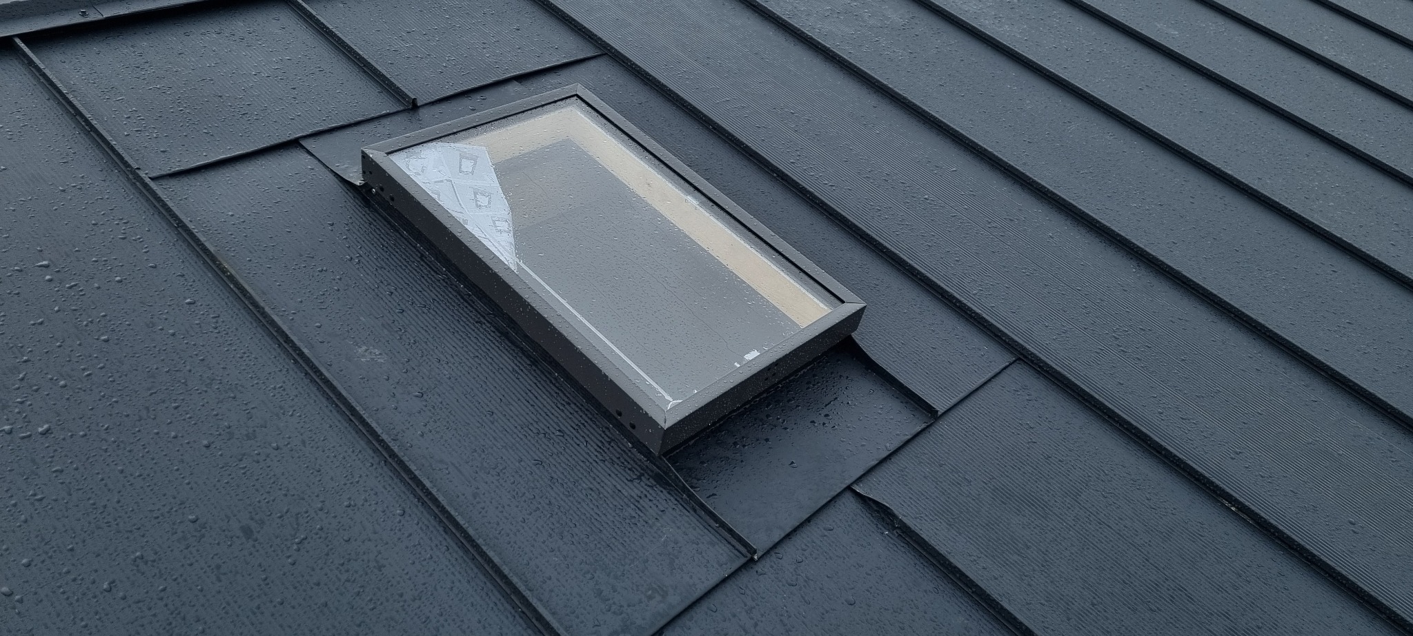 Nowoczesny dach z czarnej blachy na rąbek stojący z wbudowanym oknem dachowym, pokryty kroplami deszczu.