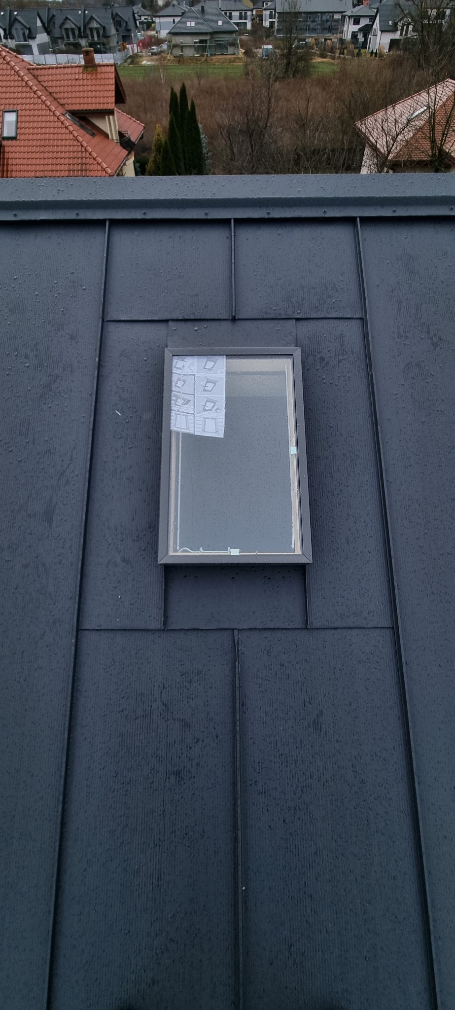 Szary dach z blachy na rąbek stojący z oknem dachowym zabezpieczonym folią montażową, widok z góry na tle zabudowy jednorodzinnej w pochmurny dzień.