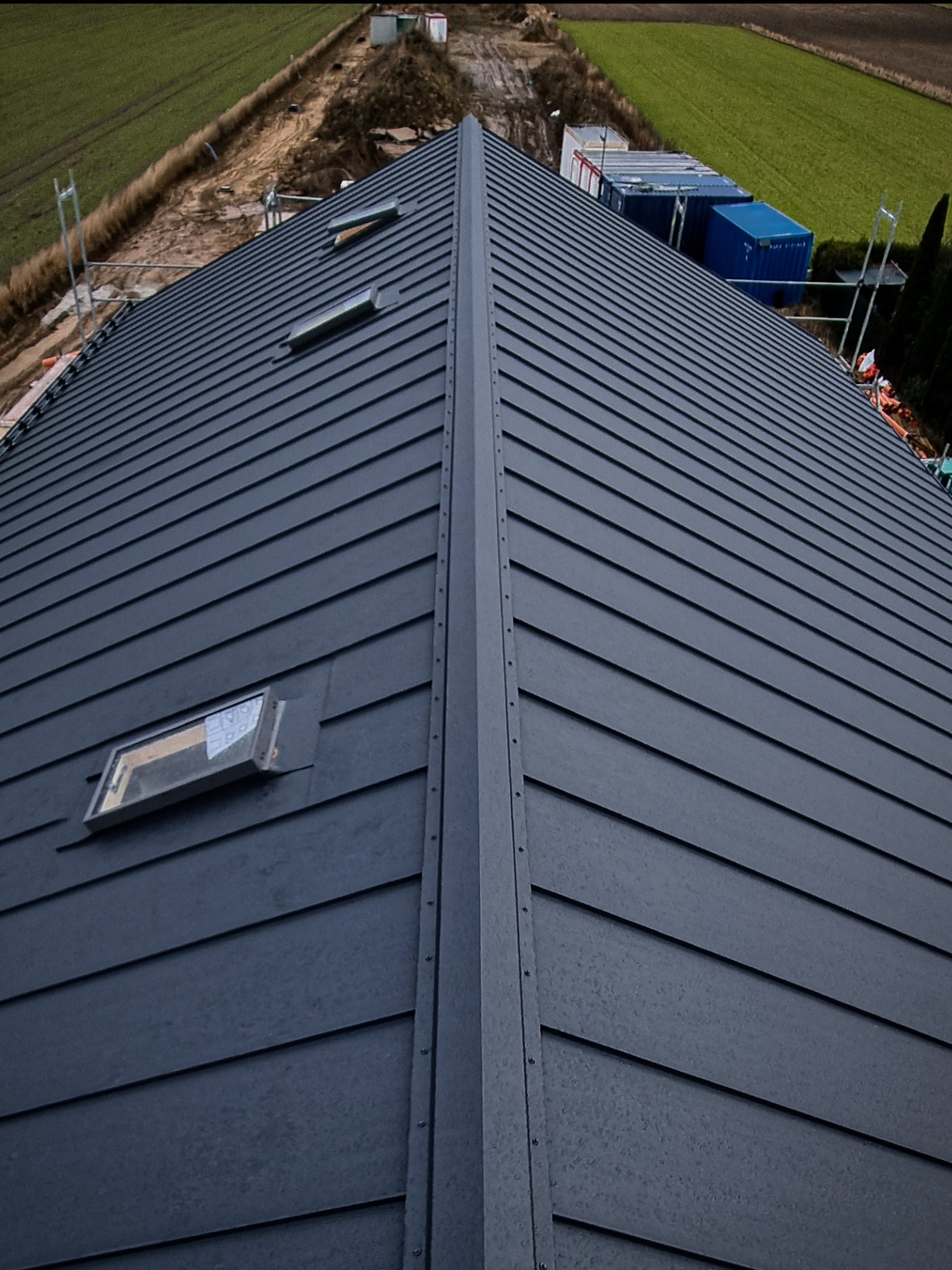 Nowoczesny, grafitowy dach z blachy na rąbek stojący z dwoma oknami dachowymi, widok z góry na tle zielonego pola i kontenerów budowlanych.