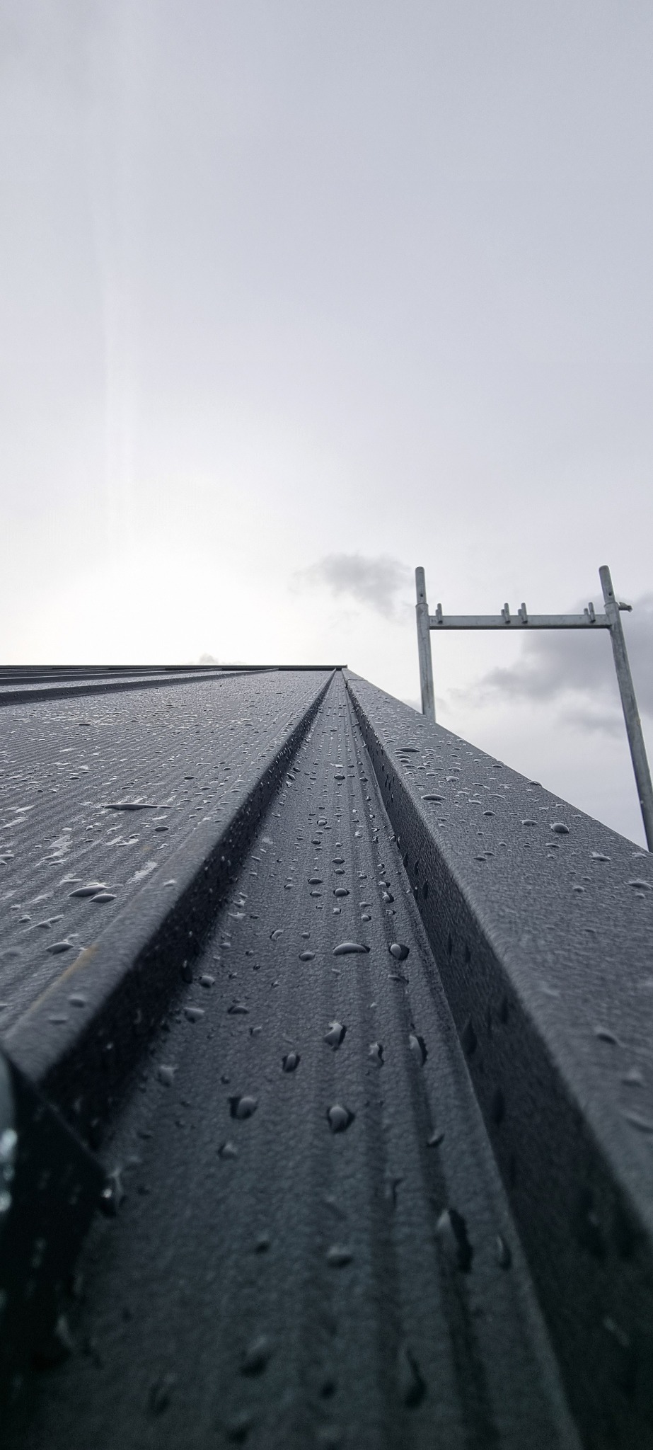 Metalowe pokrycie dachowe w kroplach deszczu, sfotografowane pod kątem, z widocznym fragmentem rusztowania i pochmurnym niebem w tle.