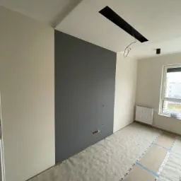 Wnętrze pokoju w trakcie malowania: jedna ściana pomalowana na szaro, reszta na kremowo, podłoga zabezpieczona folią malarską, widoczne gniazdka elektryczne i otwór w suficie z wystającymi kablami.