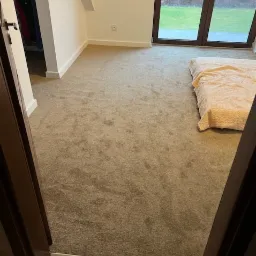 Beżowa wykładzina w pokoju z oknem na ogród, widok zza otwartych drzwi. Minimalistyczny wystrój, jasne ściany i schludny wygląd.