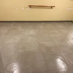 Beżowe, kwadratowe płytki podłogowe z widoczną okrągłą plamą po środku, ściany w odcieniach żółci z widocznymi śladami po usuniętych elementach. Widok z perspektywy wejścia.