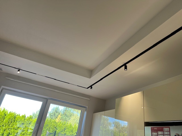 Minimalistyczne oświetlenie szynowe w kuchni z białymi szafkami. Czarne szyny z reflektorami na suficie podwieszanym, widok z dołu, okno z widokiem na zieleń.