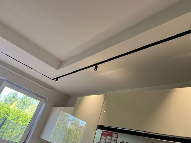 Biały sufit z czarną szyną oświetleniową i reflektorami punktowymi, widok z dołu na fragment kuchni z oknem.