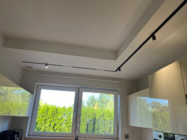 Nowoczesne oświetlenie szynowe w kuchni z jasnymi szafkami i dużym oknem z widokiem na zieleń. Czarna szyna z reflektorami na białym suficie.