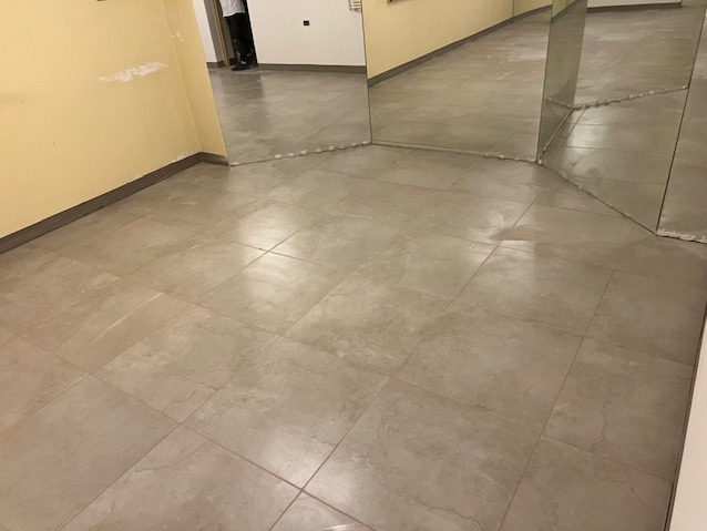 Beżowe płytki podłogowe w pokoju z lustrami i fragmentem żółtej ściany. Widoczne fugi i listwy przypodłogowe. Ujęcie z niskiej perspektywy.