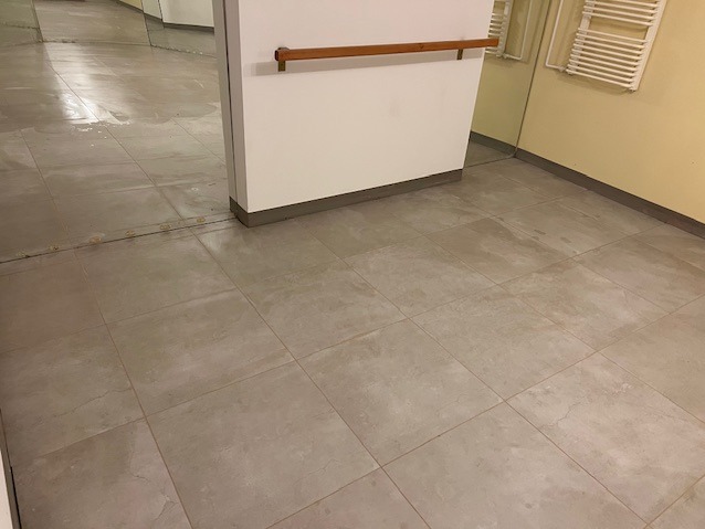 Ułożone szare płytki podłogowe w łazience, widoczne lustro i grzejnik drabinkowy. Minimalistyczny design z drewnianym akcentem.