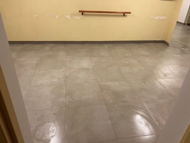 Beżowe, kwadratowe płytki podłogowe z widoczną okrągłą plamą po środku, ściany w odcieniach żółci z widocznymi śladami po usuniętych elementach. Widok z perspektywy wejścia.