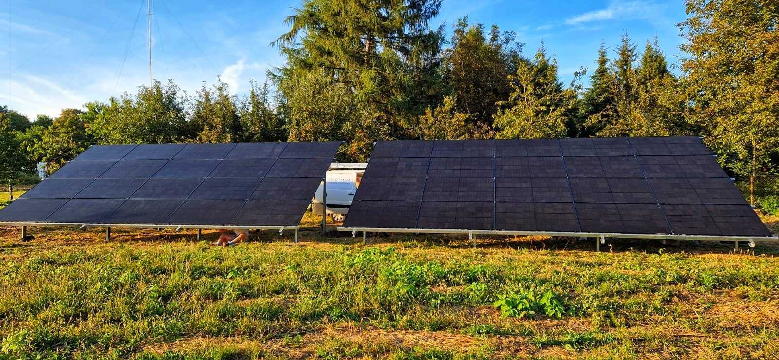 Dwa panele słoneczne ustawione na metalowej konstrukcji na trawiastym terenie, częściowo zasłaniające białego busa; w tle drzewa i maszt telekomunikacyjny.