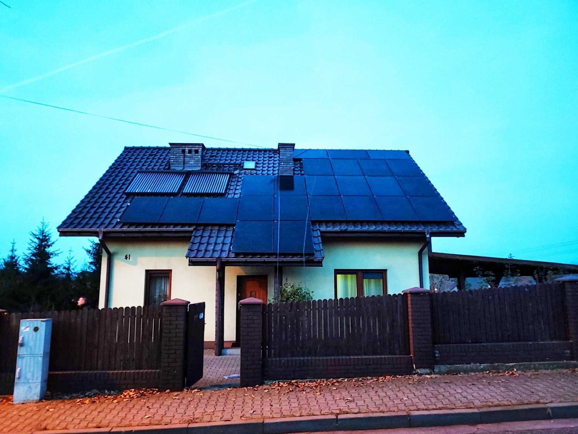 Dom jednorodzinny z brązowym dachem, na którym zamontowano panele słoneczne i kolektory słoneczne, otoczony drewnianym ogrodzeniem z ceglanymi słupkami, sfotografowany o zmierzchu.