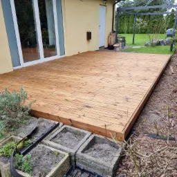 Nowy, drewniany taras przylegający do domu z jasną elewacją, widoczne betonowe donice z ziemią wzdłuż krawędzi tarasu i metalowa pergola w tle.