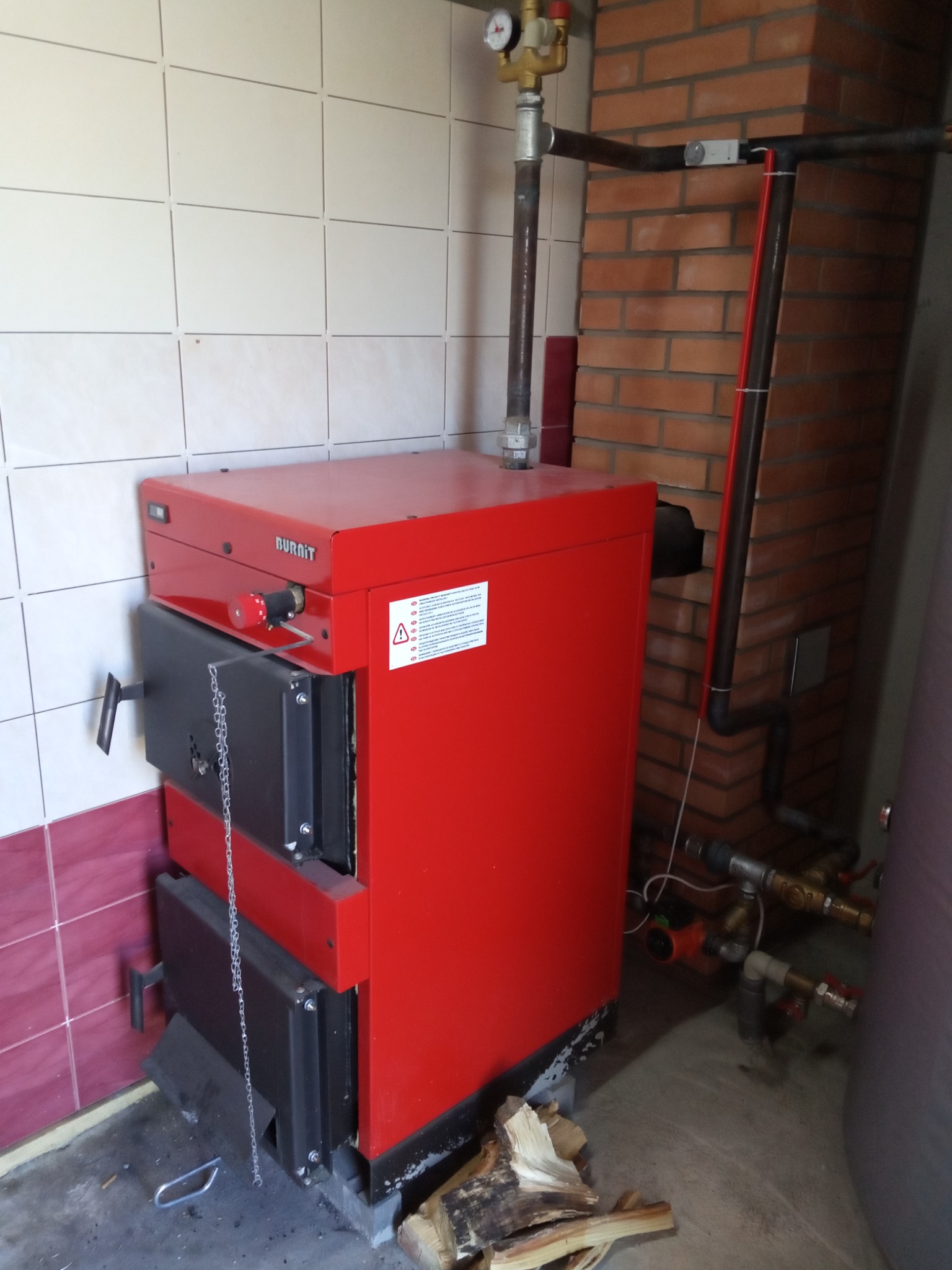 Czerwony piec centralnego ogrzewania marki Burnit, z dwoma czarnymi drzwiczkami i łańcuchem, umieszczony w kotłowni z częściowo wykafelkowanymi ścianami i widocznymi rurami instalacji grzewczej.