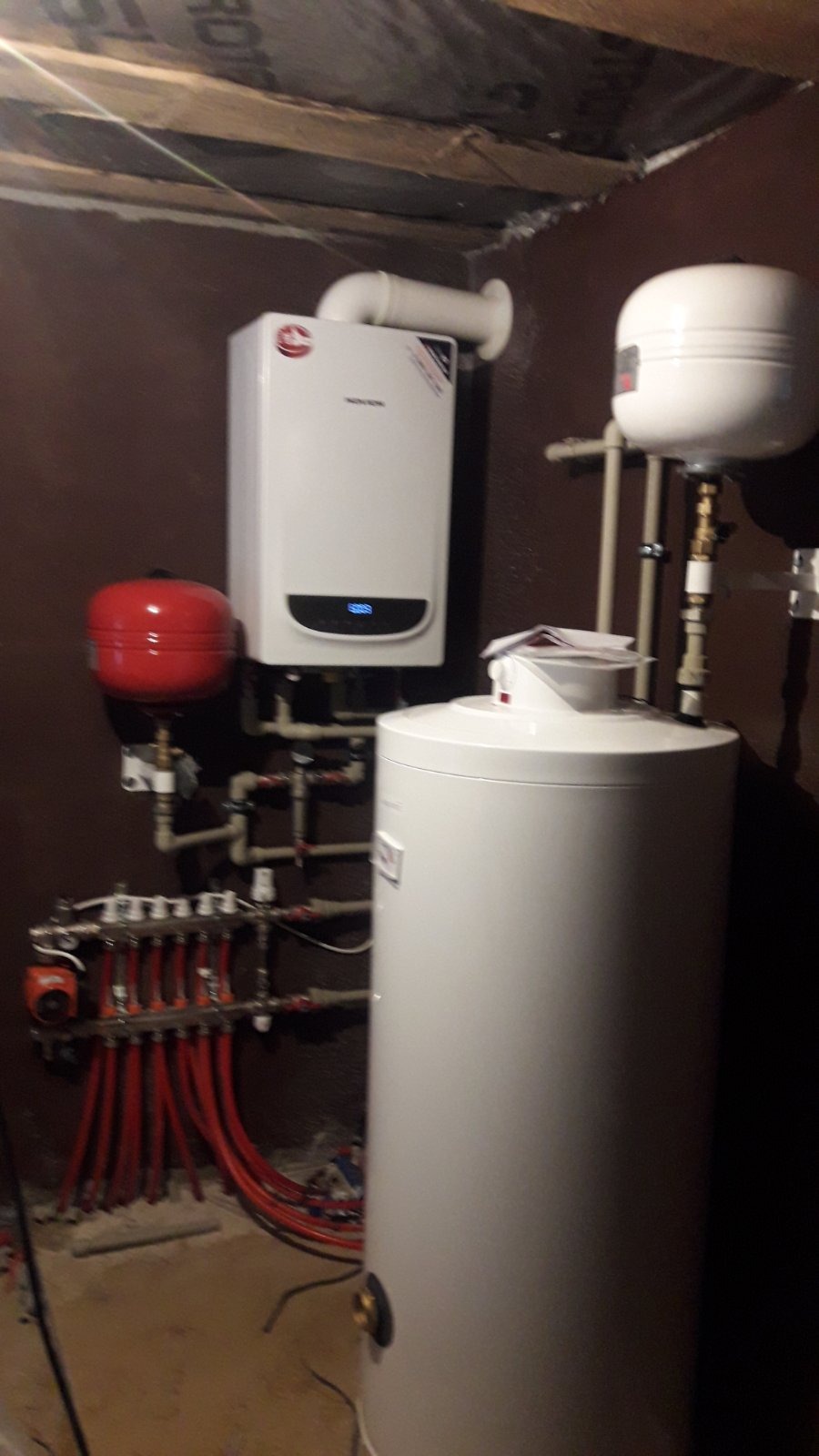 Kotłownia z zamontowanym piecem gazowym, zasobnikiem ciepłej wody, czerwonym naczyniem przeponowym i rozdzielaczem ogrzewania podłogowego z czerwonymi rurami, widok w rogu pomieszczenia z widoczną...