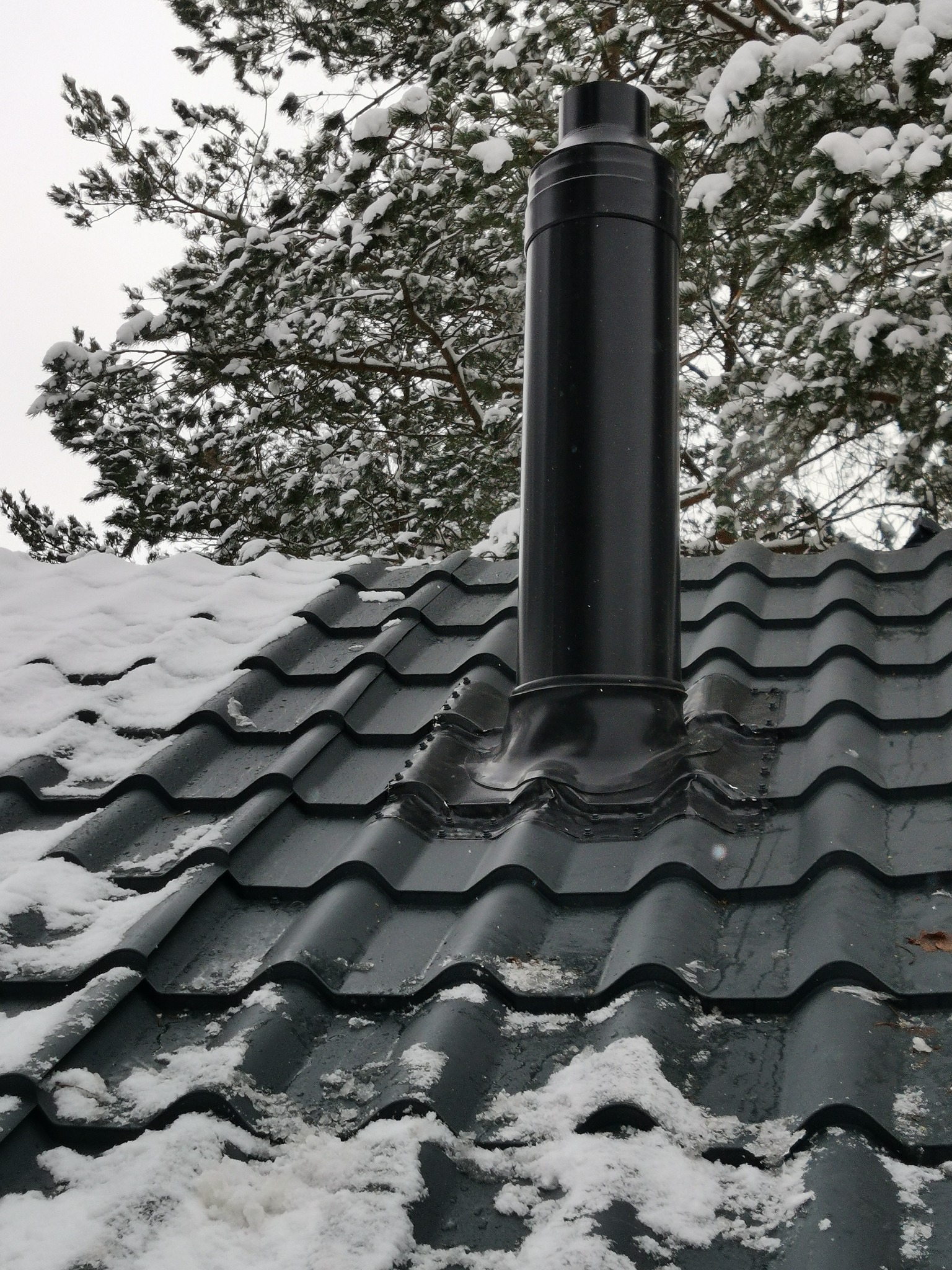 Czarny komin wentylacyjny wystający z szarego dachu pokrytego śniegiem, na tle ośnieżonego drzewa iglastego.