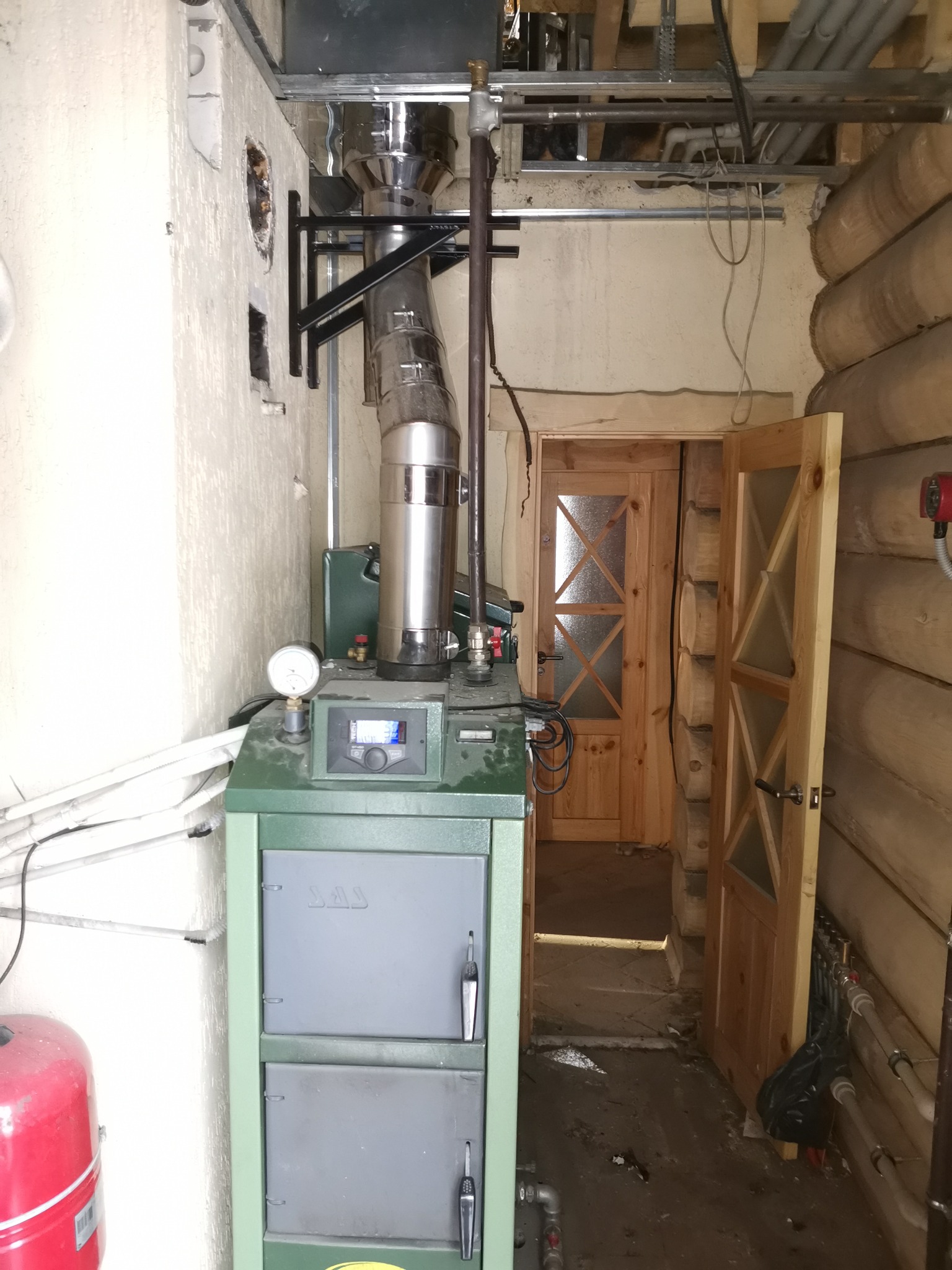 Zielony piec CO z podłączonym kominem i instalacją w pomieszczeniu z drewnianą ścianą i otwartymi drzwiami.