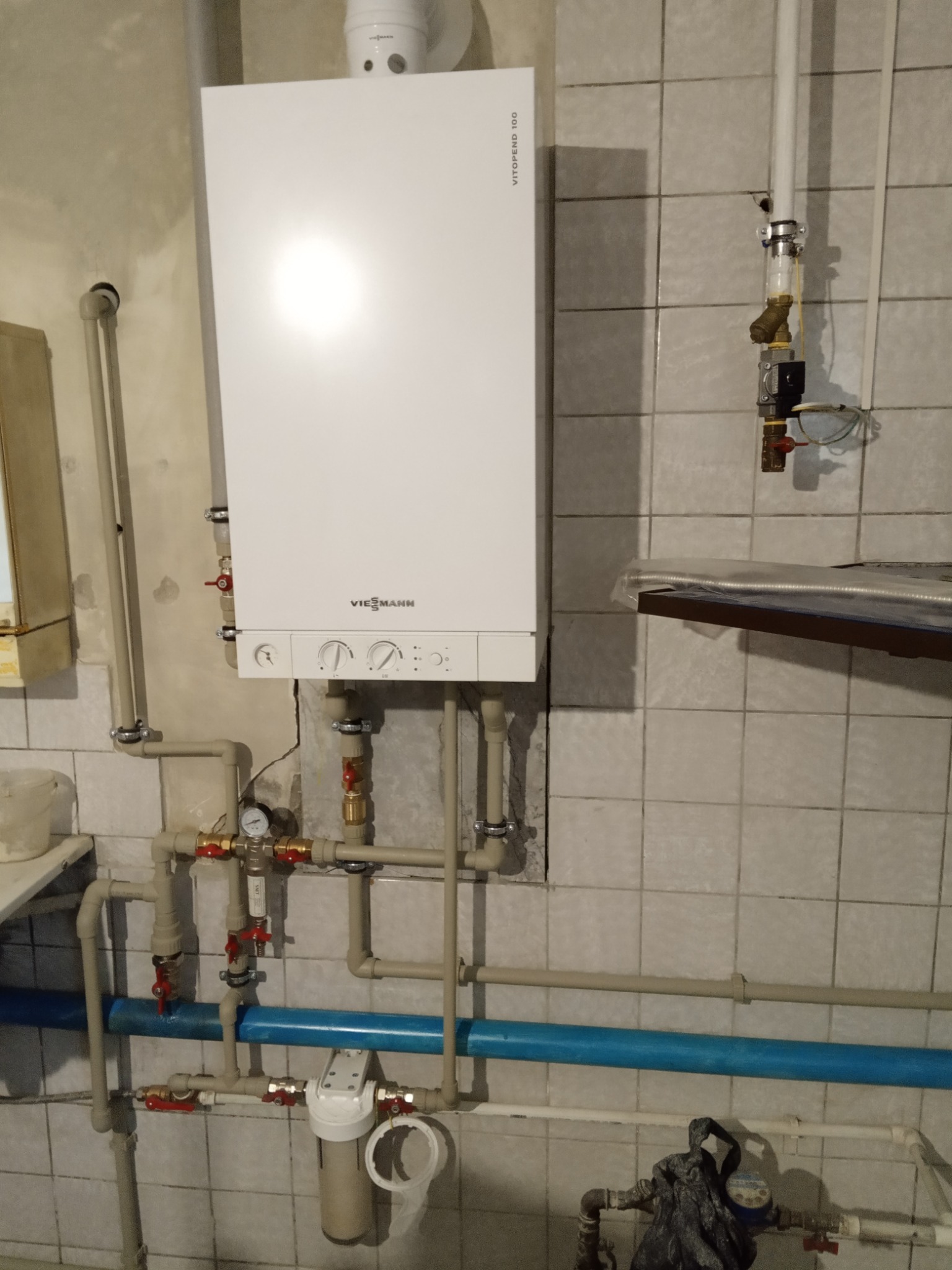 Biały piec gazowy Viessmann Vitopend 100 zamontowany na ścianie z beżowych płytek, widoczne rury doprowadzające wodę i gaz, filtr wody oraz wodomierz.