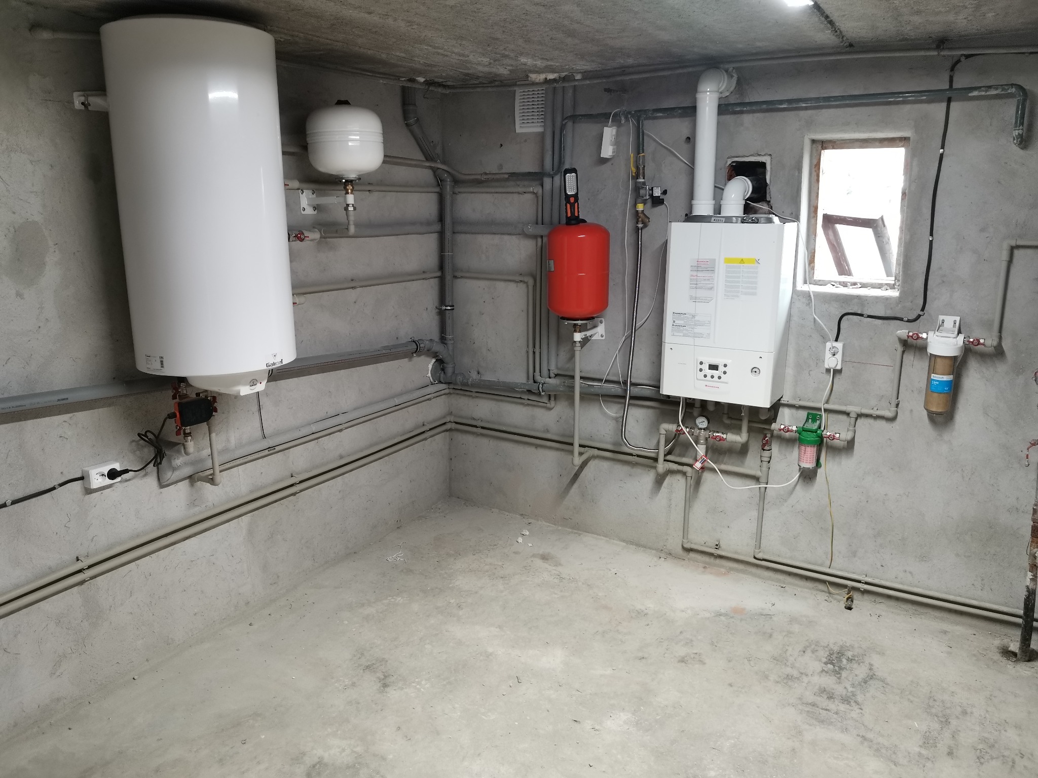 Kotłownia z zainstalowanym piecem gazowym, zasobnikiem ciepłej wody, naczyniami wzbiorczymi i filtrami wody na tle surowych betonowych ścian i podłogi.