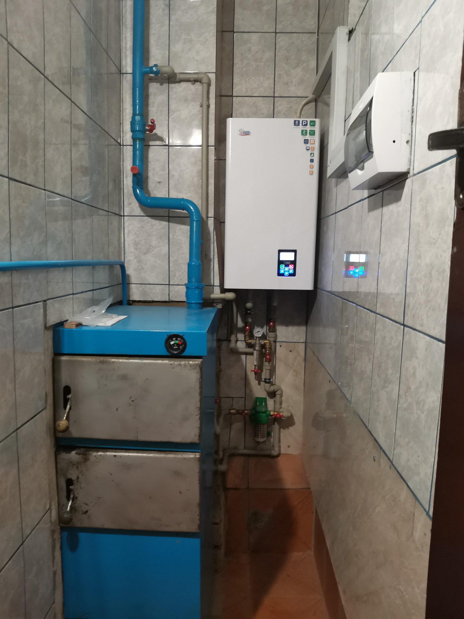 Kocioł grzewczy gazowy zamontowany na ścianie w pomieszczeniu z wyłożonymi kafelkami, obok stoi piec z niebieską obudową i srebrnymi drzwiczkami, widoczne rury i zawory.