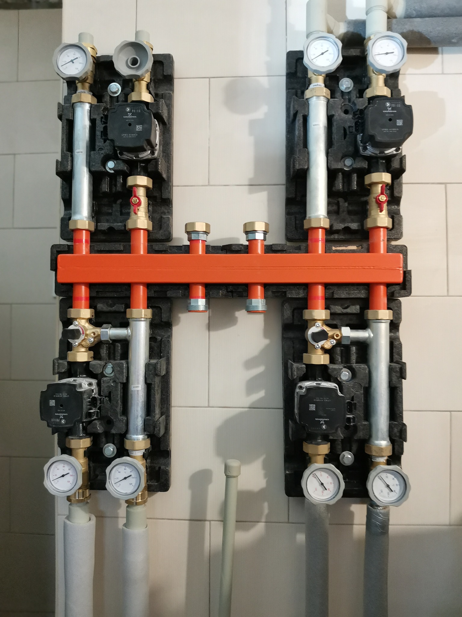 Rozdzielacz ogrzewania podłogowego z pompami obiegowymi, manometrami i rurami w izolacji, zamontowany na ścianie z płytek ceramicznych.