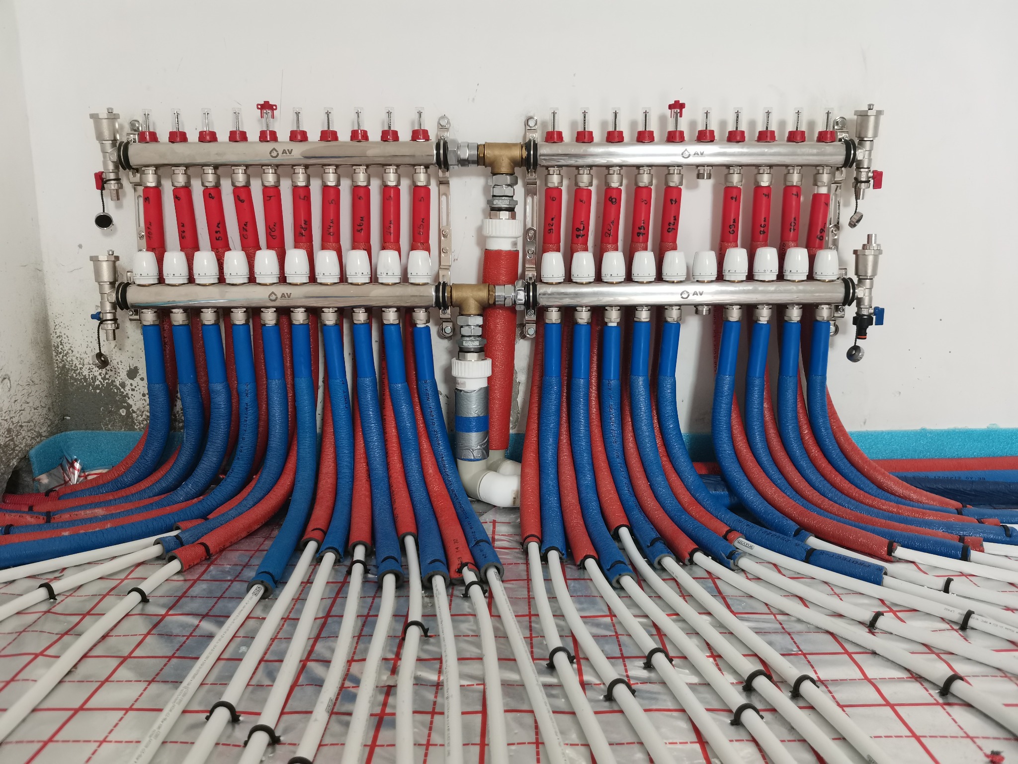 Rozdzielacz ogrzewania podłogowego z licznymi pętlami rur w kolorach czerwonym i niebieskim, ułożonymi na siatce grzewczej, widok z góry, na ścianie zarys instalacji.