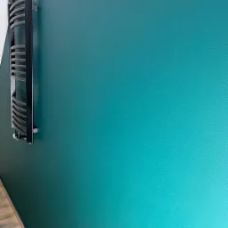 Ściana w odcieniach turkusu z zamontowanym, nowoczesnym grzejnikiem drabinkowym z czarną ramą i szarymi szczeblami, widok z boku.