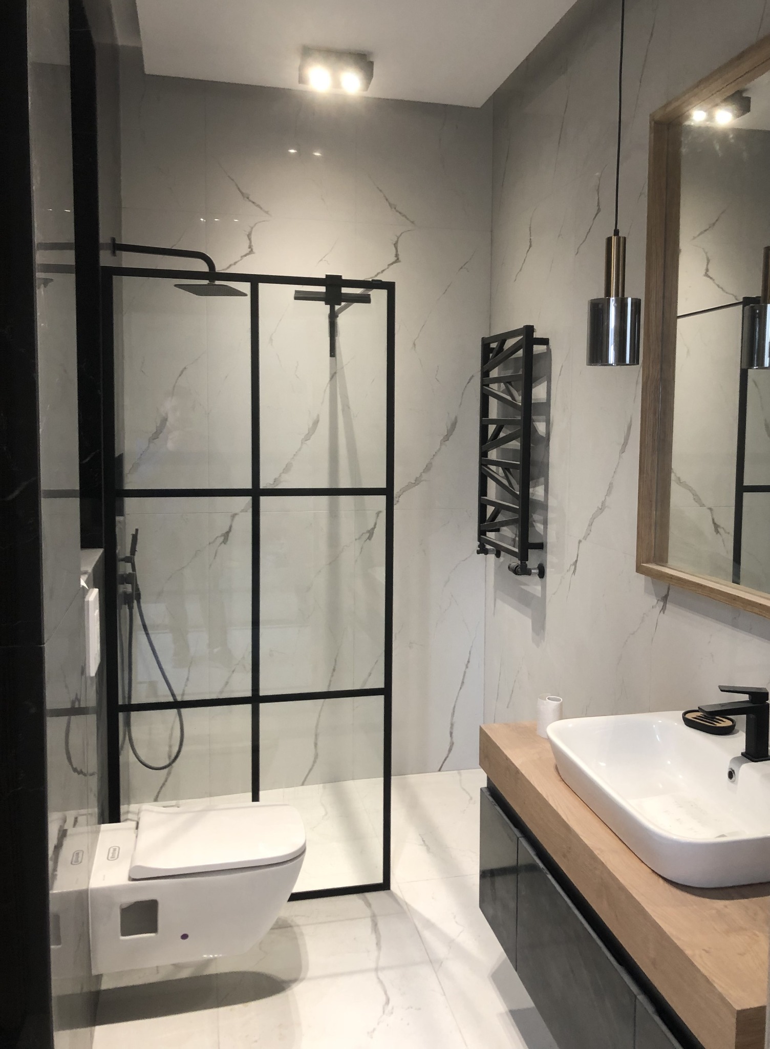 Nowoczesna łazienka z kabiną prysznicową typu walk-in z czarnymi ramami, wiszącą toaletą, umywalką nablatową na drewnianym blacie i grzejnikiem drabinkowym w kolorze czarnym.