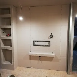 Wnętrze w trakcie prac wykończeniowych z płyt kartonowo-gipsowych: zabudowa ściany z wnękami, przygotowane miejsce na telewizor i półka, widoczne profile metalowe i oświetlenie budowlane.